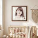 A framed portrait print of a young girl with brown hair hangs on a beige wall above a light wood crib in a softly styled nursery. The print has a dark brown frame, and the room includes a macrame wall hanging, bedding, and a small stuffed toy. - Thumbnail 2