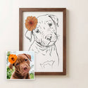 A framed custom pet portrait print featuring a black line art sketch of a puppy with an orange flower accent, shown next to a small reference photo of a brown puppy on a light beige wall. - Thumbnail 1