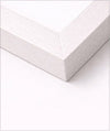 A close-up of a white picture frame corner showing the light wood grain texture of the frame and the white mat board inside, set against a clean white background.